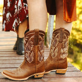 A pair of cowgirl boots with a desert clay color, featuring a snip toe and a heel, placed on a wooden surface with a natural setting in the background.