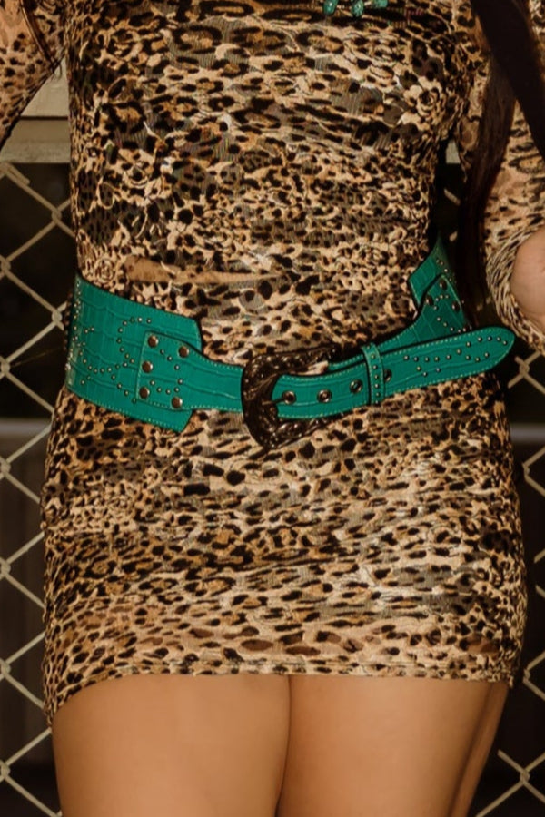 Woman in a patterned dress and high boots standing in an urban setting with a chain-link fence and concrete wall.