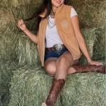 Woman sitting on a hay bale wearing a tan vest, white shirt, blue shorts, and brown boots.