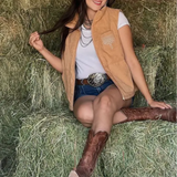 Woman sitting on a hay bale wearing a tan vest, white shirt, blue shorts, and brown boots.