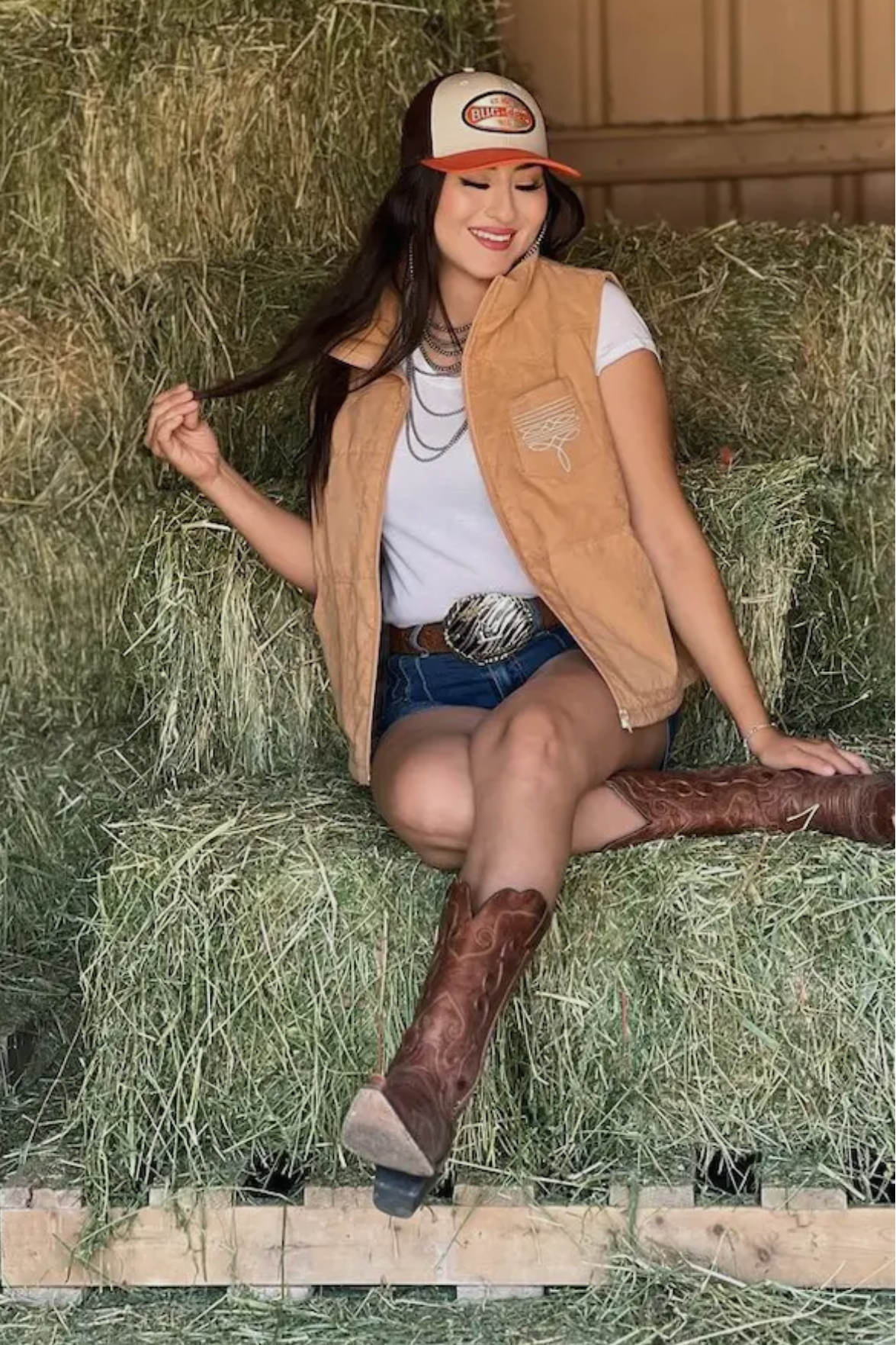 Woman sitting on a hay bale wearing a tan vest, white shirt, blue shorts, and brown boots.