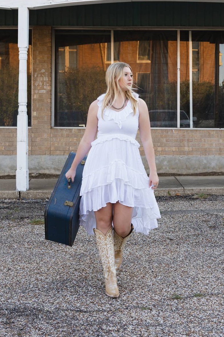 Woman in a white dress holding a blue suitcase in front of an old furniture store.