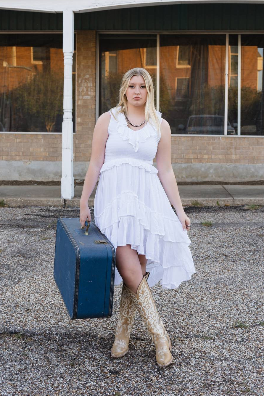 Woman in a white dress holding a blue suitcase in front of an old building with 'INS FUR' sign.