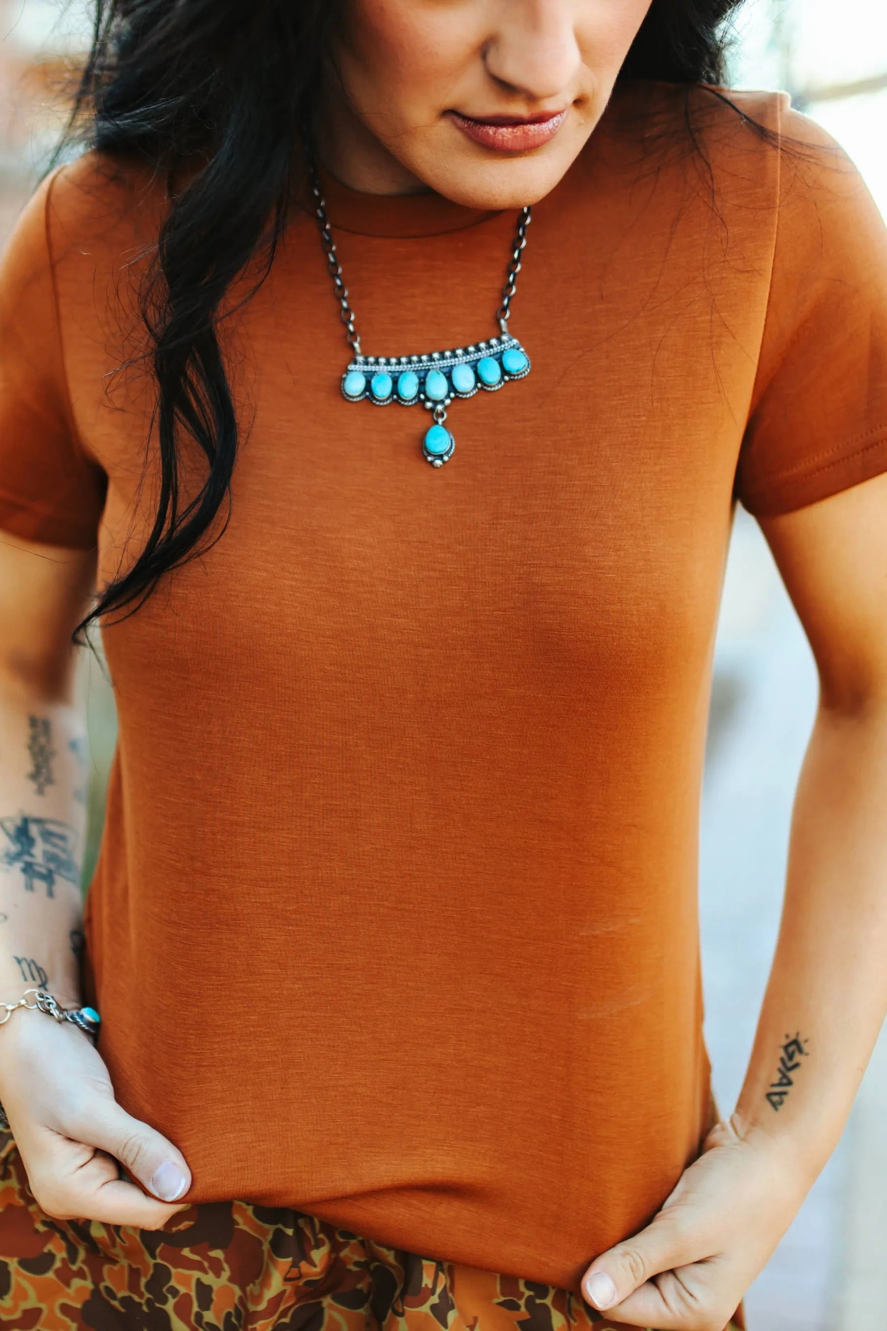 A person wearing a brown fitted t-shirt with a decorative necklace and a patterned skirt.