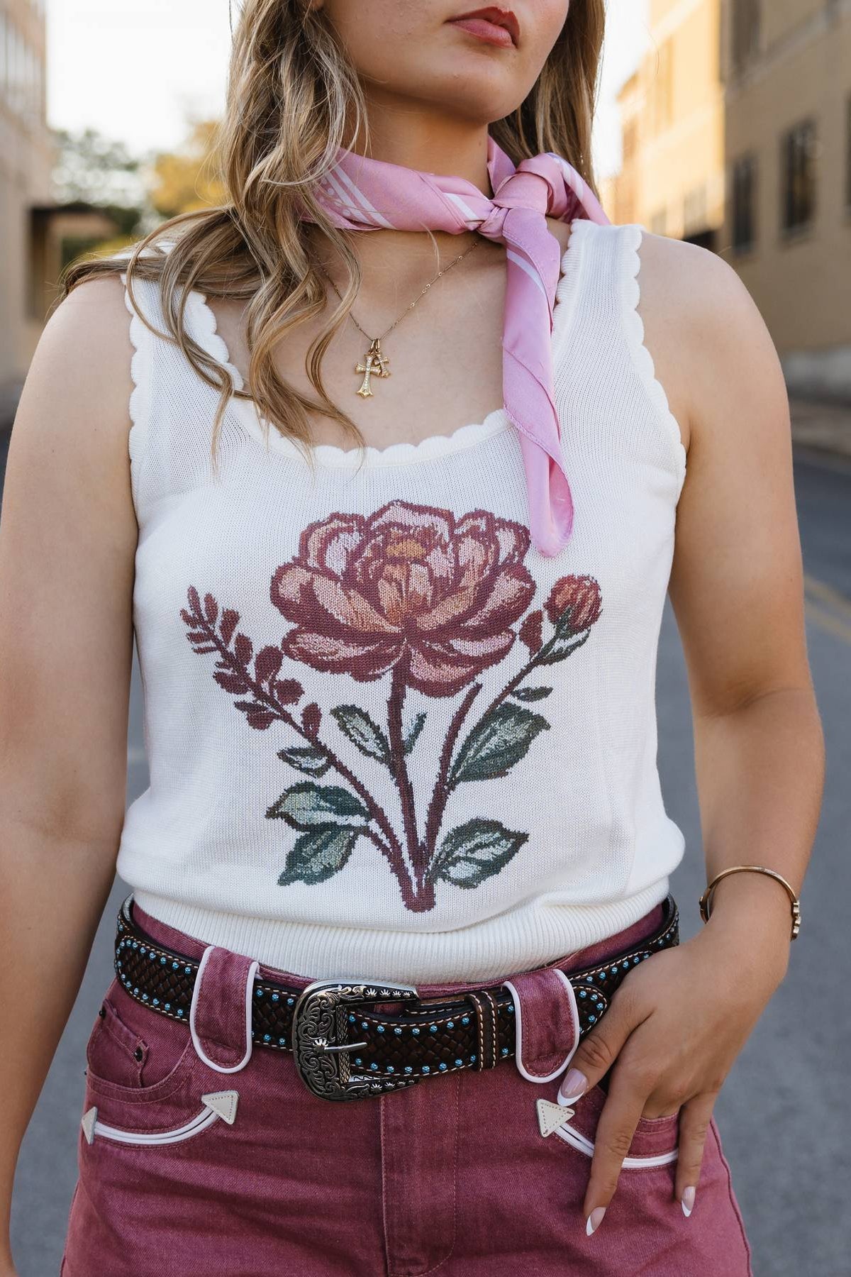 Person wearing a white tank top with a floral design, pink scarf, and maroon pants on a street.