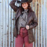 Person wearing a cowboy hat and brown leather jacket against a rusty corrugated metal wall