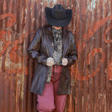 Person wearing a cowboy hat and leather jacket standing against a rusty corrugated metal wall with 'Coca-Cola' branding.