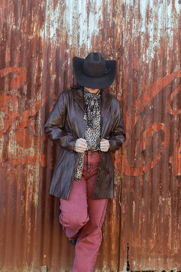 Person wearing a cowboy hat and leather jacket standing against a rusty corrugated metal wall with 'Coca-Cola' branding.