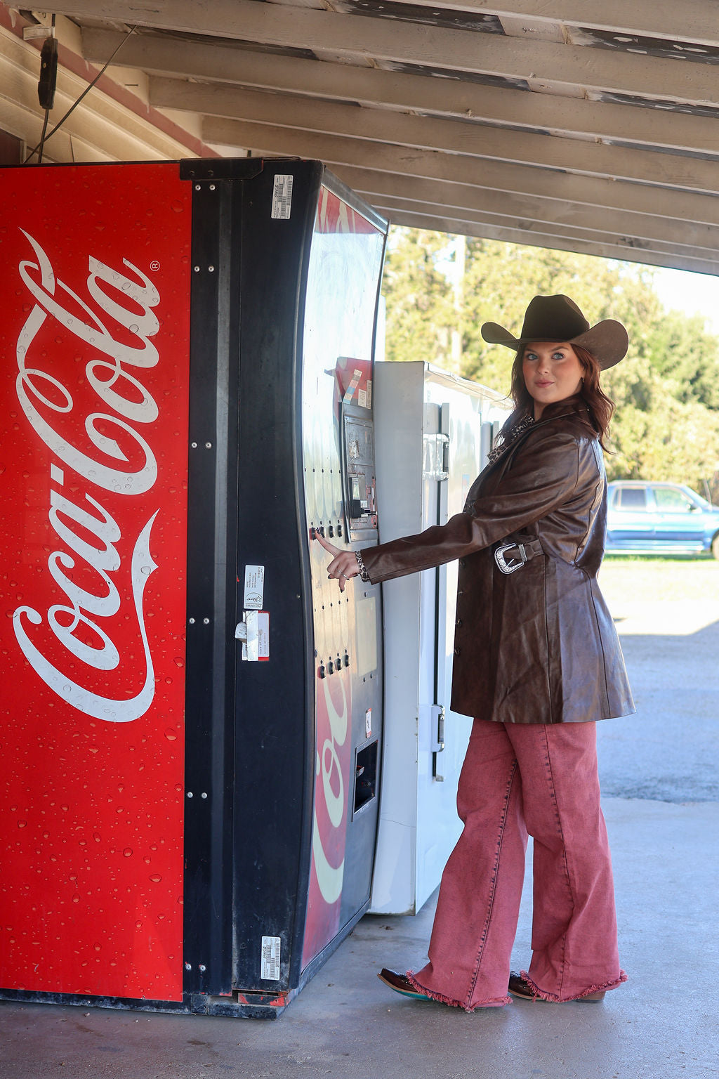 Person in cowboy hat and leather jacket standing next to a Coca-Cola vending machine.