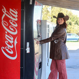 Person in cowboy hat and leather jacket standing next to a Coca-Cola vending machine.