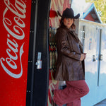 Person in cowboy hat and leather jacket standing next to a Coca-Cola vending machine.