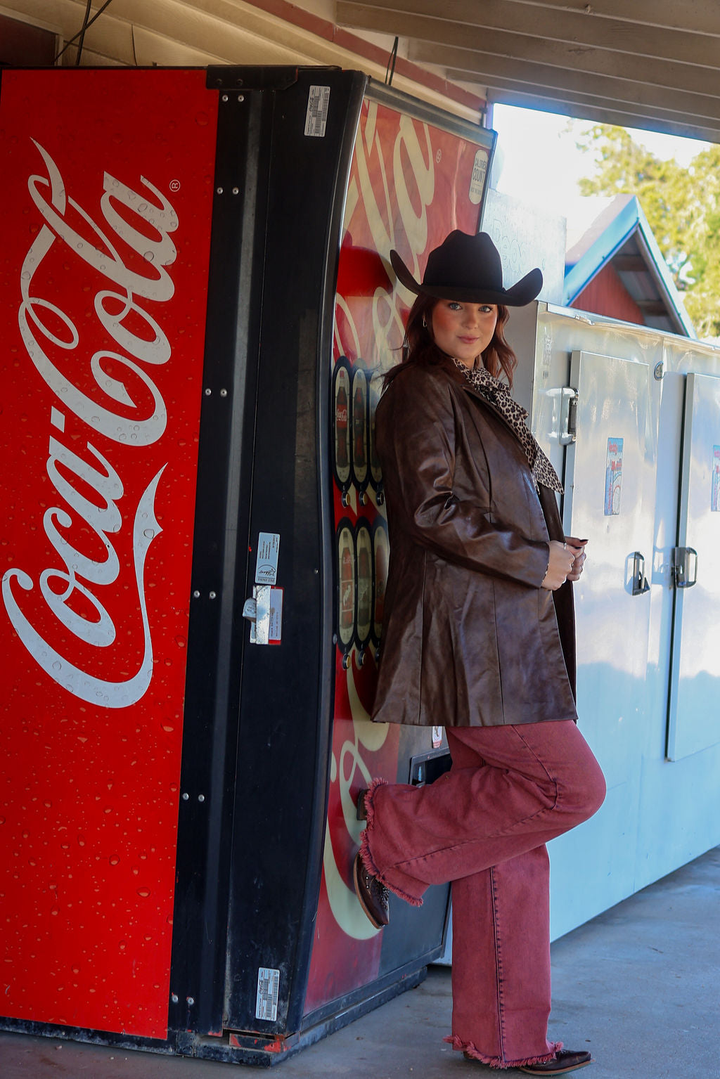 Person in cowboy hat and leather jacket standing next to a Coca-Cola vending machine.