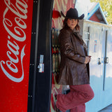 Person in cowboy hat and leather jacket standing next to a Coca-Cola vending machine.