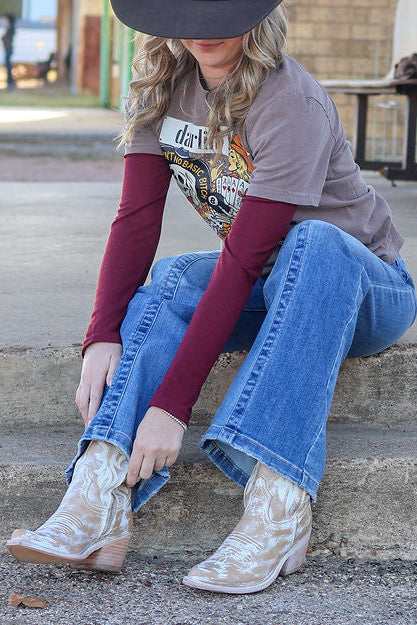 Person sitting on steps wearing a cowboy hat, jeans, and boots in front of a small building.