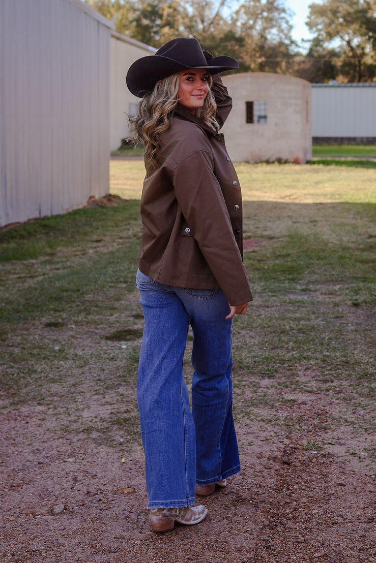 Person wearing a cowboy hat, brown jacket, and blue jeans standing on a dirt path with trees and buildings in the background.