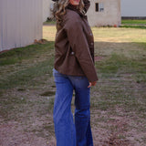 Person wearing a cowboy hat, brown jacket, and blue jeans standing on a dirt path with trees and buildings in the background.