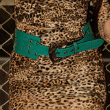 Woman in a patterned dress and high boots standing in an urban setting with a chain-link fence and concrete wall.